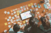 3 people sitting over a table with the Conceptboard online collaboration tool being open in front of them. There are many sticky notes spread on the table, but with conceptoboard you can brainstorm directly on the board