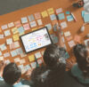 3 people sitting over a table with the Conceptboard online collaboration tool being open in front of them. There are many sticky notes spread on the table, but with conceptoboard you can brainstorm directly on the board