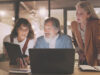 3 people from project management team working together. One person has a laptop in the hand, while the middle person has a laptop on the table