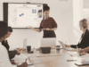 Eine Frau steht in einem Konferenzraum vor einem interaktiven Whiteboard, auf dem Remote-Teilnehmende zugeschaltet sind.